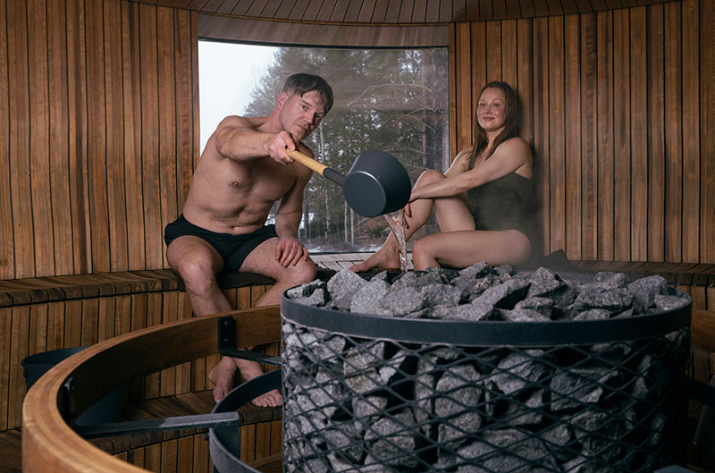 Mężczyzna polewa chochlą wodę na rozgrzane kamienie sauny, obok siedzi kobieta; okrągła, parująca drewniana sauna z oknem.